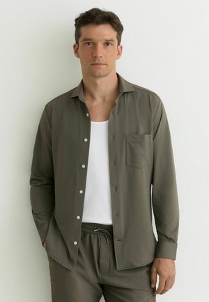 Noor mees seisab valge seina ees, kandes oliivrohelise tekstuuriga nööpidega särki valge tanktopi ja sobivate pükstega.