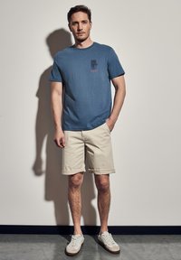 Modré bavlněné tričko s krátkým rukávem, výsoké s malým grafickým potiskem. V kombinaci s béžovými šortkami s ohrnutými nohavicemi a světlými teniskami.