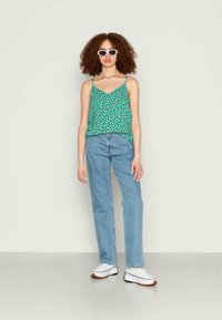 Mulher com cabelo encaracolado a usar óculos de sol brancos, um top floral verde, jeans azul claros e ténis brancos, de pé contra um fundo neutro.