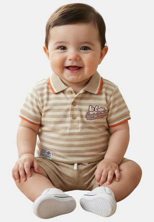 Bébé souriant aux cheveux foncés, portant un polo beige à rayures, un short assorti et des chaussures blanches, assis sur un fond blanc.