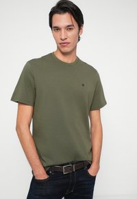 Ung mann iført olivengrønn Calvin Klein t-skjorte med lite logo, mørke jeans og brun belte, stående med hendene i lommer.