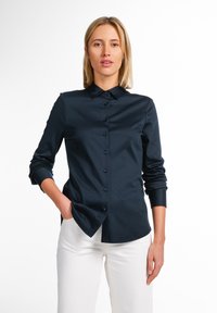 Camicia button-up a maniche lunghe blu navy con colletto classico, dotata di bottoni frontali e una texture morbida e liscia. Abbinata a pantaloni bianchi.