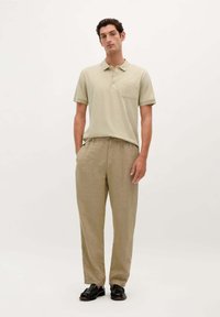 Camisa polo color beige claro con un bolsillo en el pecho, combinada con pantalones de lino beige de corte holgado, complementados con zapatos negros. Diseño minimalista.