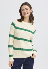 Pull crème à rayures horizontales vertes, texture tricotée, encolure ronde et poignets côtelés. Associé à un jean bleu foncé.