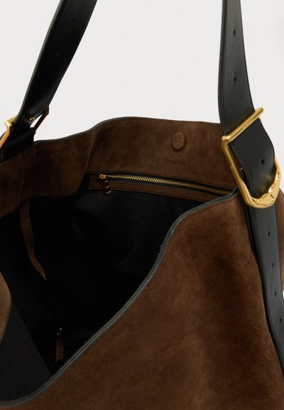 Sacola de camurça castanha com uma alça de couro preta, apresentando um bolso com fecho dourado e um interior liso. Design minimalista e textura suave.