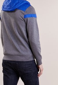 Felpa grigia con cappuccio in tessuto trapuntato blu e accenti sulle spalle; design aderente, polsini e vita a coste. Texture morbida, stile casual.