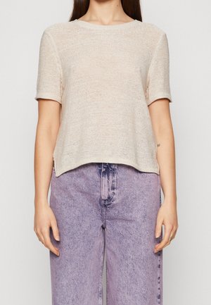 Kobieta ubrana w beżowy, krótki sweter z dzianiny i wysokowalne, szerokie, wyblakłe fioletowe dżinsy, stojąca na białym tle.