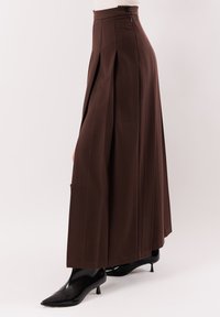 Pantaloni marroni a vita alta, a gamba larga e plissettati, abbinati a stivaletti neri con punta a punta e tacco basso, su sfondo bianco.