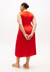 Κόκκινο πλισέ φόρεμα με στενή μπούστα και φουσκωτή φούστα, με στρογγυλή πλάτη, φορεμένο με κεντημένα καφέ σανδάλια.