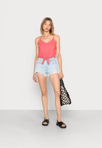 Top vermelho sem mangas com detalhes em botão e faixa na cintura, combinado com calções de ganga claros e esfarrapados e sandálias pretas. Segura uma bolsa de rede preta.