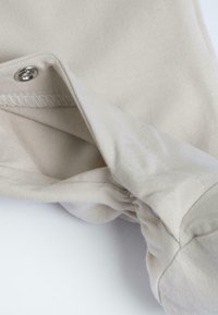 Tissu en coton beige avec une texture lisse, comportant un bouton-pression argenté, des coutures visibles, et un détail de poignet replié.