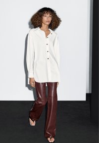 Bílá košile s dlouhým rukávem a zlatými knoflíky, doplněná o vysoce střižené kožené kalhoty ve vínově červené barvě. Látka vypadá hladce se slabým leskem.