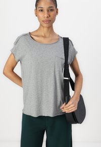 Sivý krátky rukáv tričko s výstrihom a prednou vreckom. Nosené s zelenými nohavicami. Čierna crossbody taška s vyrazeným logom.