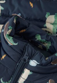 Ύφασμα μπουφάν πούφερ ναυτικού με σχέδια δεινοσαύρων και αστραπών. Διαθέτει ψηλό γιακά και μπροστινό φερμουάρ με τράβηγμα.