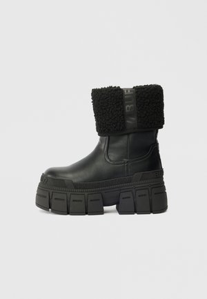 Bota de tornozelo preta em couro com sola de plataforma grossa e punho de pelo dobrável rotulada "BUFFALO" na tira e na sola.