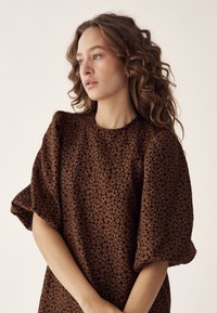 Tânără cu păr creț purtând o rochie maro cu un mic model floral, privindu-se gânditoare în lateral, pe un fundal simplu.