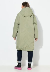 Lange olijfgroene parka met capuchon, elastische zoom en zijzakken, gecombineerd met donkere jeans en roze, dikke zool laarzen.
