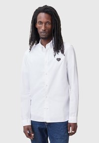 Camisa blanca de botones hecha de algodón, con un bordado de corazón negro texturizado en el lado izquierdo del pecho. Cuello clásico y mangas largas.