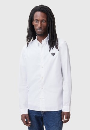 Camisa blanca de botones hecha de algodón, con un bordado de corazón negro texturizado en el lado izquierdo del pecho. Cuello clásico y mangas largas.