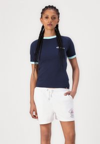 Camicia a maniche corte blu navy con polsini azzurri e logo sul petto, abbinata a pantaloni corti bianchi con design grafico e vita con cordoncino.
