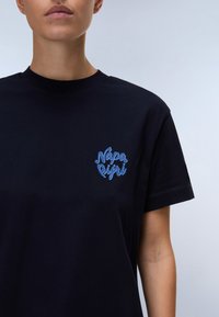 Osoba w czarnej koszulce z niebieskim logo "Nápapijri" na lewej piersi, na jednolitym jasnym tle.