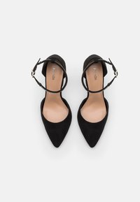 Une paire de ballerines noires en daim à bout pointu avec des brides autour de la cheville et des fermoirs en métal argenté, présentées sur un fond blanc.