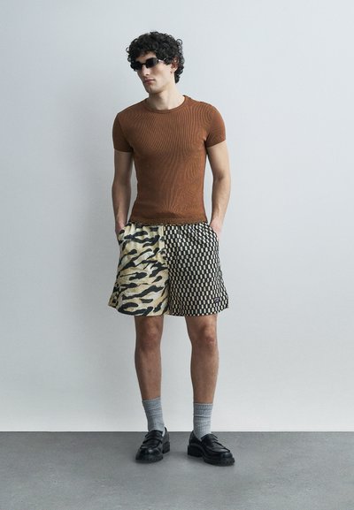 Άνδρας που φοράει καφέ εφαρμοστή μπλούζα, μισό με animal print και μισό καρό σορτς, γκρι κάλτσες, μαύρα παπούτσια και μαύρα γυαλιά ηλίου, στέκεται μπροστά σε ένα γκρι τοίχο.