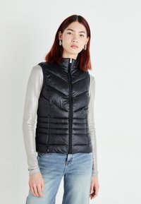 Black, puffy sleeveless vest with vertical and chevron quilting, zip closure, and high collar, worn over a gray long-sleeve top and blue jeans.
