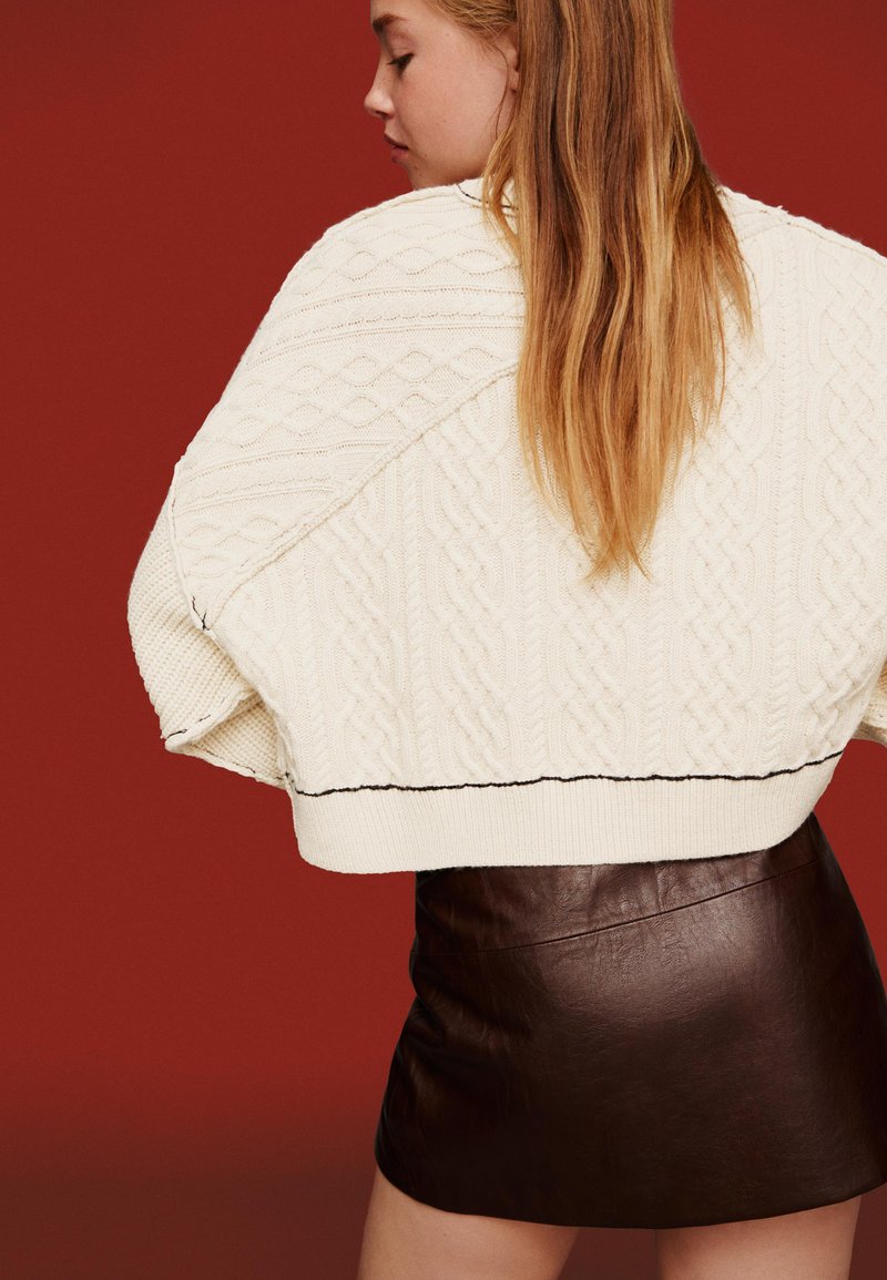 Vitt stickat tröja med flätade mönster och svart accentstygning, kombinerad med en mörkbrun läder minikjol mot en röd bakgrund.