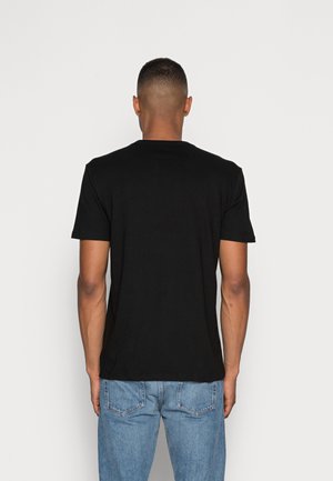 Mężczyzna o krótkich włosach, ubrany w jednolity czarny, krótki rękaw T-shirt i niebieskie dżinsy, pokazany od tyłu na jednolitym białym tle.