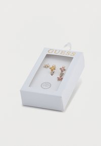 Σκουλαρίκια σε σχήμα λουλουδιού με χρυσαφένιο τόνο μέσα σε κουτί· διαθέτουν διάφανα, κίτρινα και ροζ πολύτιμους λίθους, βάση από ανοξείδωτο ατσάλι και λευκή υφασμάτινη συσκευασία.