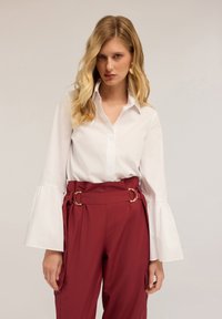 Blouse blanche à manches évasées, associée à un pantalon bordeaux taille haute avec des accents en cercle doré et une coupe lisse et ajustée.