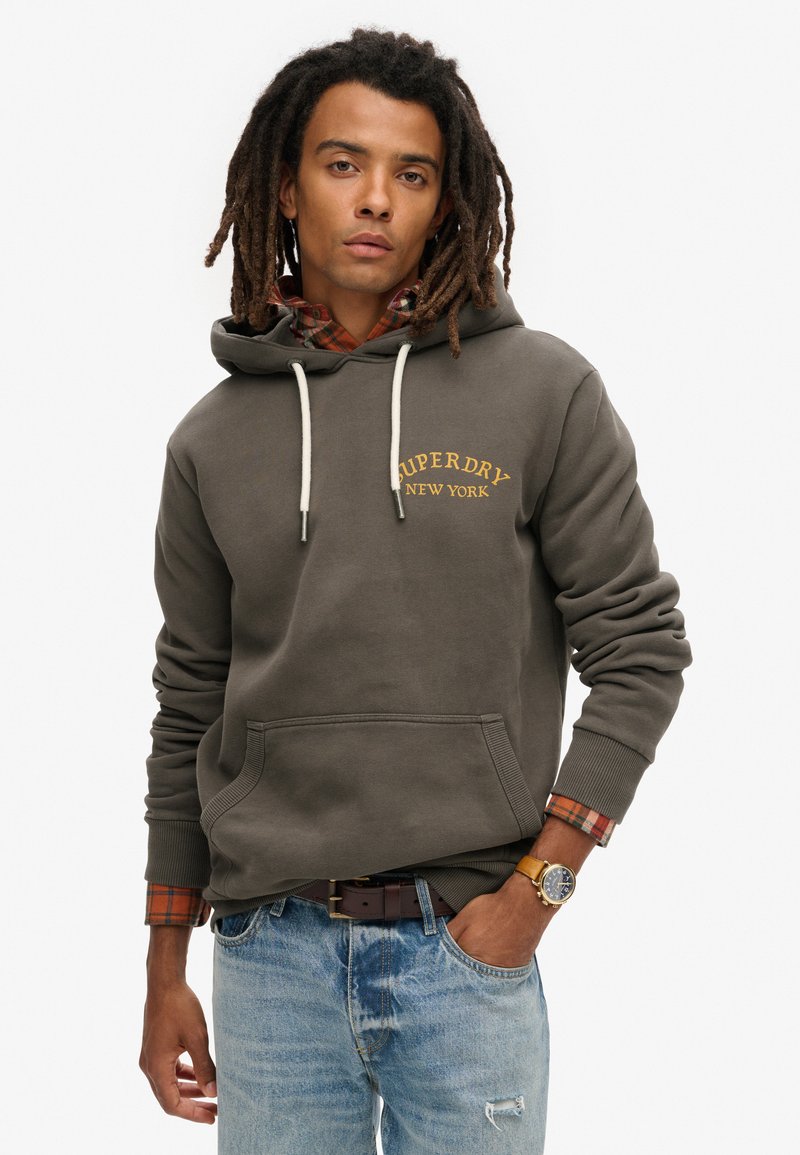 Olijfgroene hoodie met koord, voorzak en 'SUPERDRY NEW YORK' logo. Draag het over een rood en zwart geruite shirt en lichtblauwe jeans.