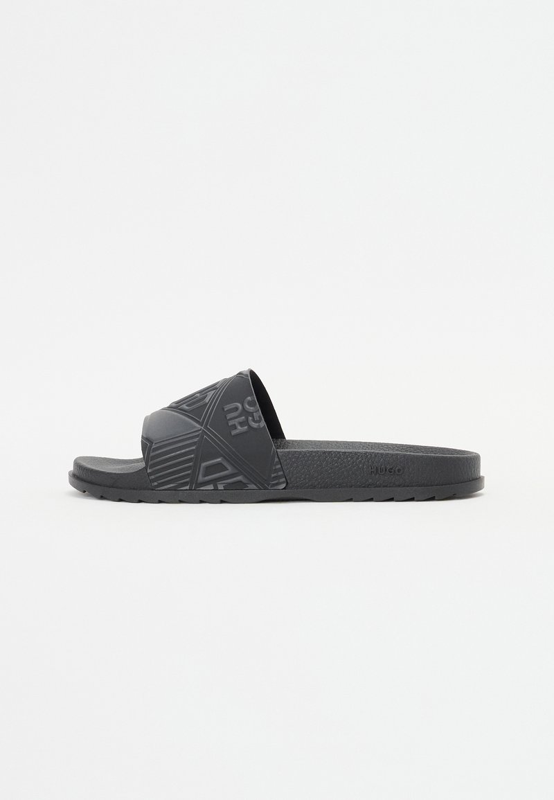Crne gumene klizne sandale s širokim remenom koji ima teksturirani geometrijski uzorak i utisnuti logo. Ravni đon s teksturiranim gripom.