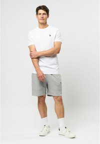 Balti medvilninė T-shirt, pilki šortai su logotipu ir balti sportbačiai. Modelis stovi su sukryžiuotomis rankomis ant vienspalvės fono.