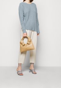 Beżowa skórzana torebka z unikanym węzłowym uchwytem, noszona z jasnoniebieskim prążkowanym swetrem i beżowymi zwężającymi się spodniami. Sandały na obcasie w niebieskim kolorze.