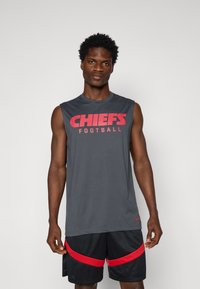 Pilkas be rankovių marškinėliai su raižytu raudonu užrašu "CHIEFS FOOTBALL"; derinami su juodais šortais su raudonomis detalėmis ir tekstūruotu dizainu.