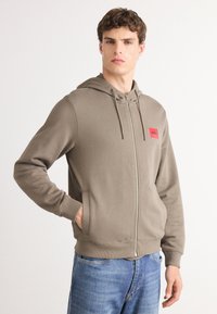Sudadera con capucha y cierre de cremallera en mezcla de algodón marrón claro, que presenta un parche de logo rojo en el lado izquierdo del pecho y puños y dobladillo de canalé.