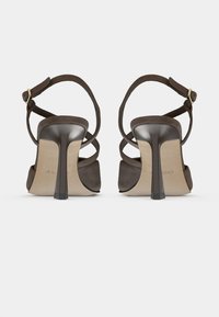 Sandales à talons hauts en daim marron avec brides à la cheville et détail croisé à l'arrière. Elles présentent un talon brillant et une semelle intérieure beige avec un logo.