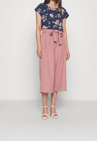 Floral navy blouse with short sleeves, paired with high-waisted, pink culottes featuring a front bow detail and wide, flowing legs. White heeled sandals.