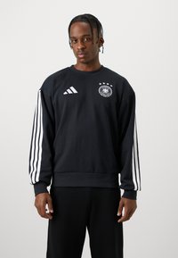 Svart hettegenser med hvite Adidas-striper på ermene og en rund logo med fire stjerner. Myk stoff, rund hals og ribbestrikkede mansjetter.