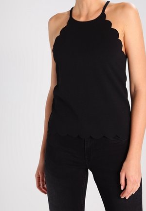 Femme portant un haut noir sans manches à bords festonnés et un pantalon noir sur un fond clair uni.