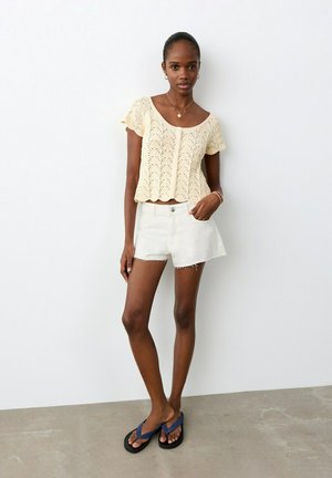 Femme debout contre un mur blanc portant un haut en maille crème à manches courtes, un short blanc taille haute et des tongs bleues.