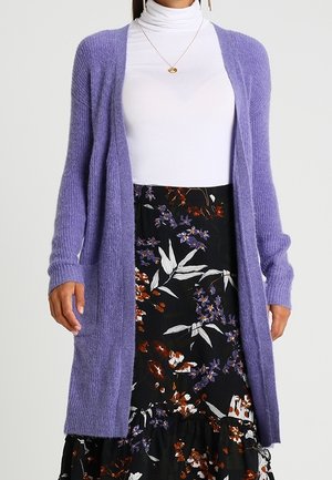 Kvinde iført en hvid rullekrave, lang lilla cardigan og sort blomsterkjole med lilla og orange blomster.