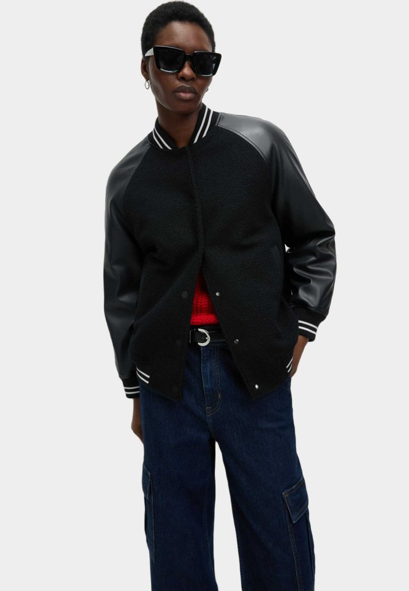 Chaqueta universitaria negra con tejido texturizado y mangas de cuero, cuello acanalado a rayas; combinada con pantalones oscuros y una camiseta roja. Se llevan gafas de sol negras.