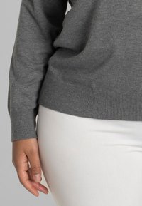 Pulover gri din tricot cu un tiv rotund, cu manșete ribbate și o textură moale. Asortat cu pantaloni de culoare crem.