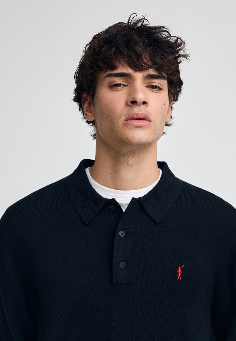 Polo shirt in maglia nera con colletto, dotato di una patta con tre bottoni e un piccolo emblema rosso sul lato sinistro del petto. Tessuto testurizzato.