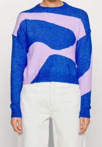 Person som bär en blå och lavendelfärgad tröja med abstrakt mönster, långa ärmar och hög midja, med armarna avslappnat längs sidorna.