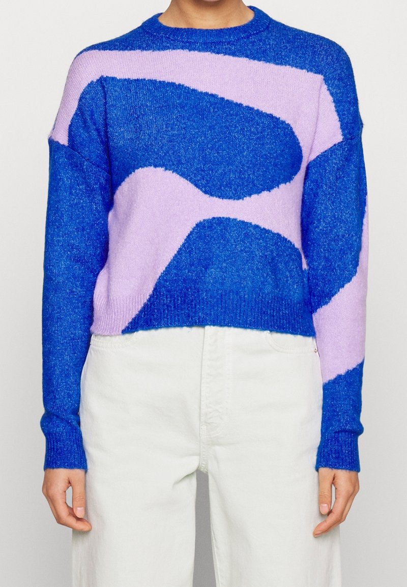 Person som bär en blå och lavendelfärgad tröja med abstrakt mönster, långa ärmar och hög midja, med armarna avslappnat längs sidorna.
