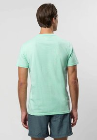 Miętowozielona koszulka z krótkim rękawem i okrągłym dekoltem, wykonana z miękkiego materiału. Noszona z wzorzystymi szortami, widok od tyłu.